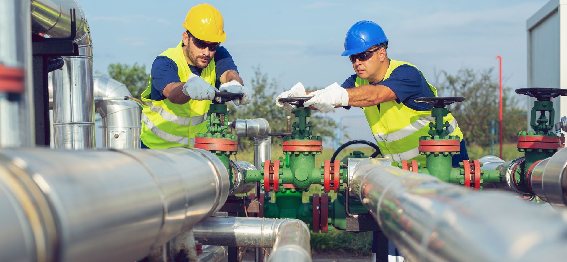 Two workers in high-visibility vests and hard hats turning industrial valves at an outdoor facility.