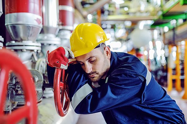 A worker in a yellow hard hat and blue uniform leans exhausted against a large red industrial valve in a facility.