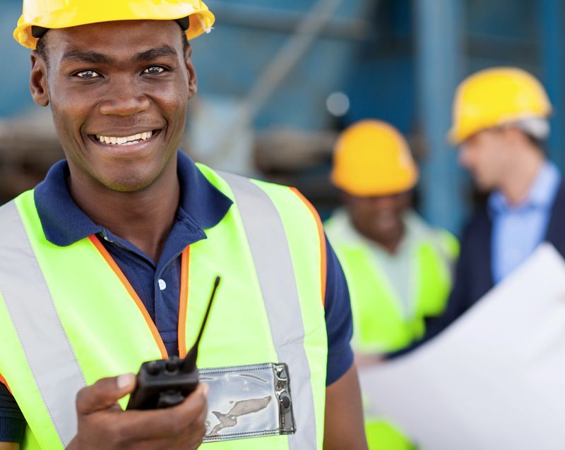 A construction worker in a high-visibility vest and yellow hard hat smiles while holding a walkie-talkie on a job site.