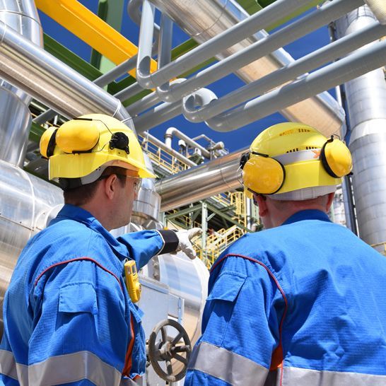 Two workers in blue uniforms and yellow hard hats inspect industrial equipment at a facility.