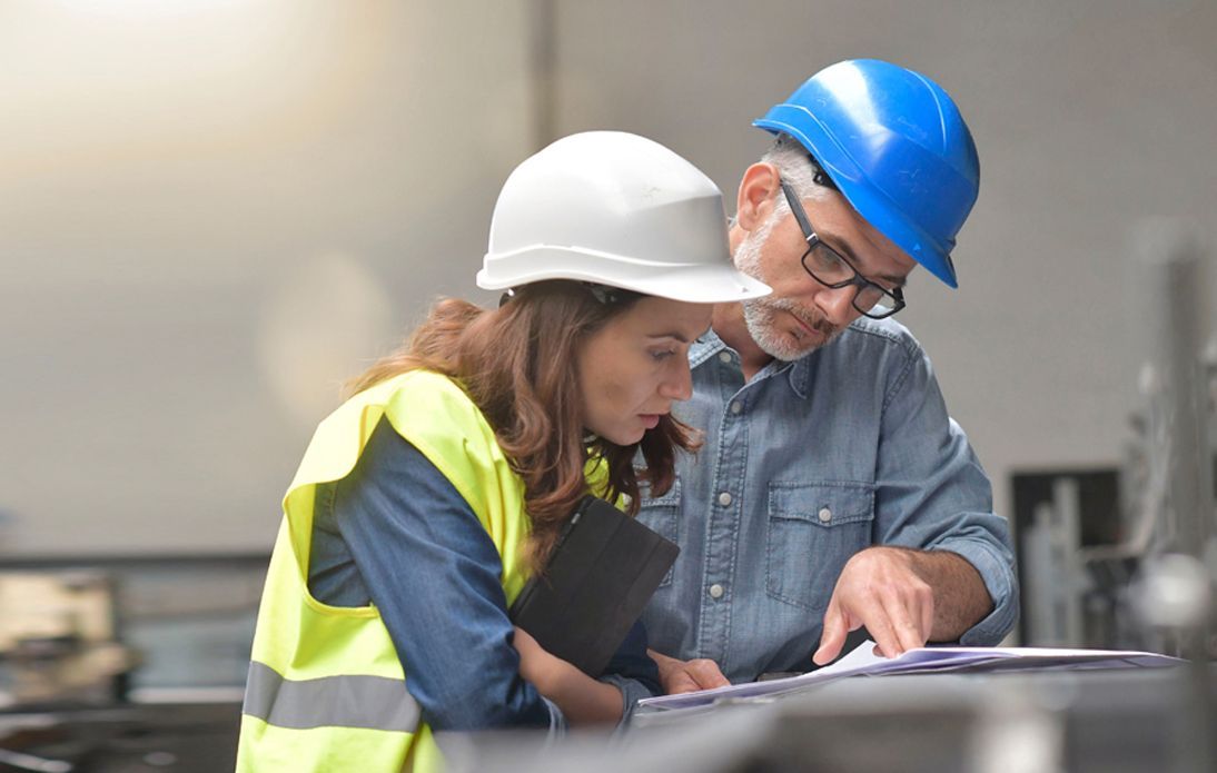 Two people in hard hats and reflective safety gear review documents together in a professional industrial workspace.