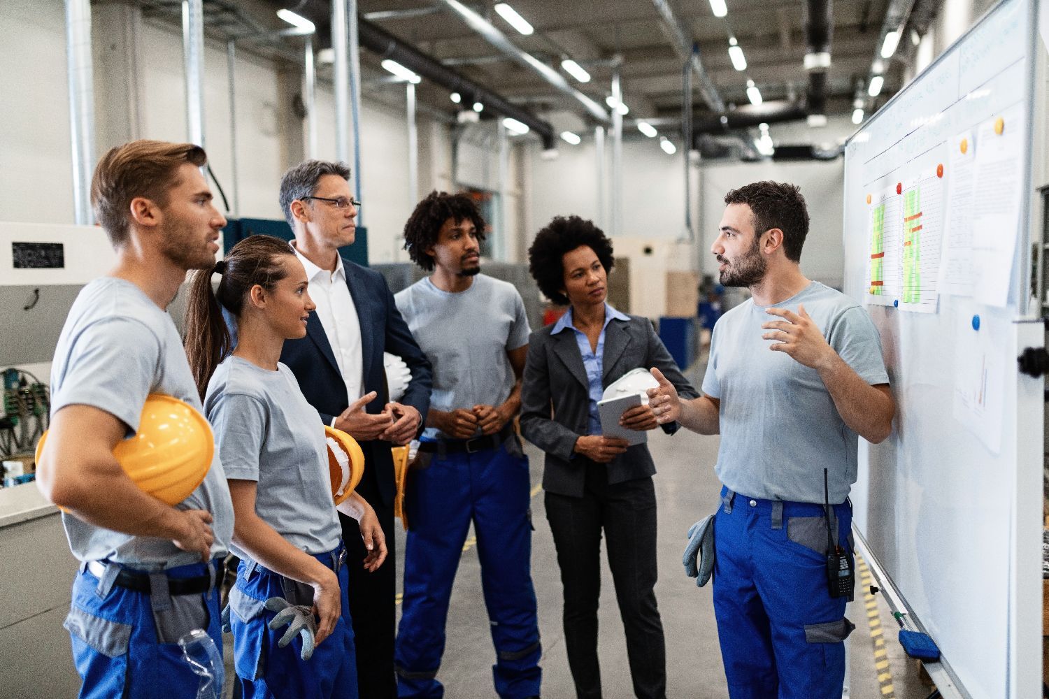 A team of workers and professionals in a manufacturing facility discuss a project plan displayed on a whiteboard.