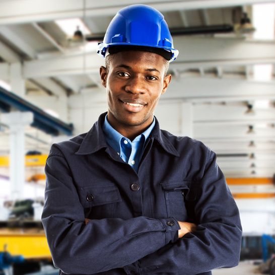 A smiling professional in a blue hard hat and uniform with arms crossed, standing in an industrial workshop.