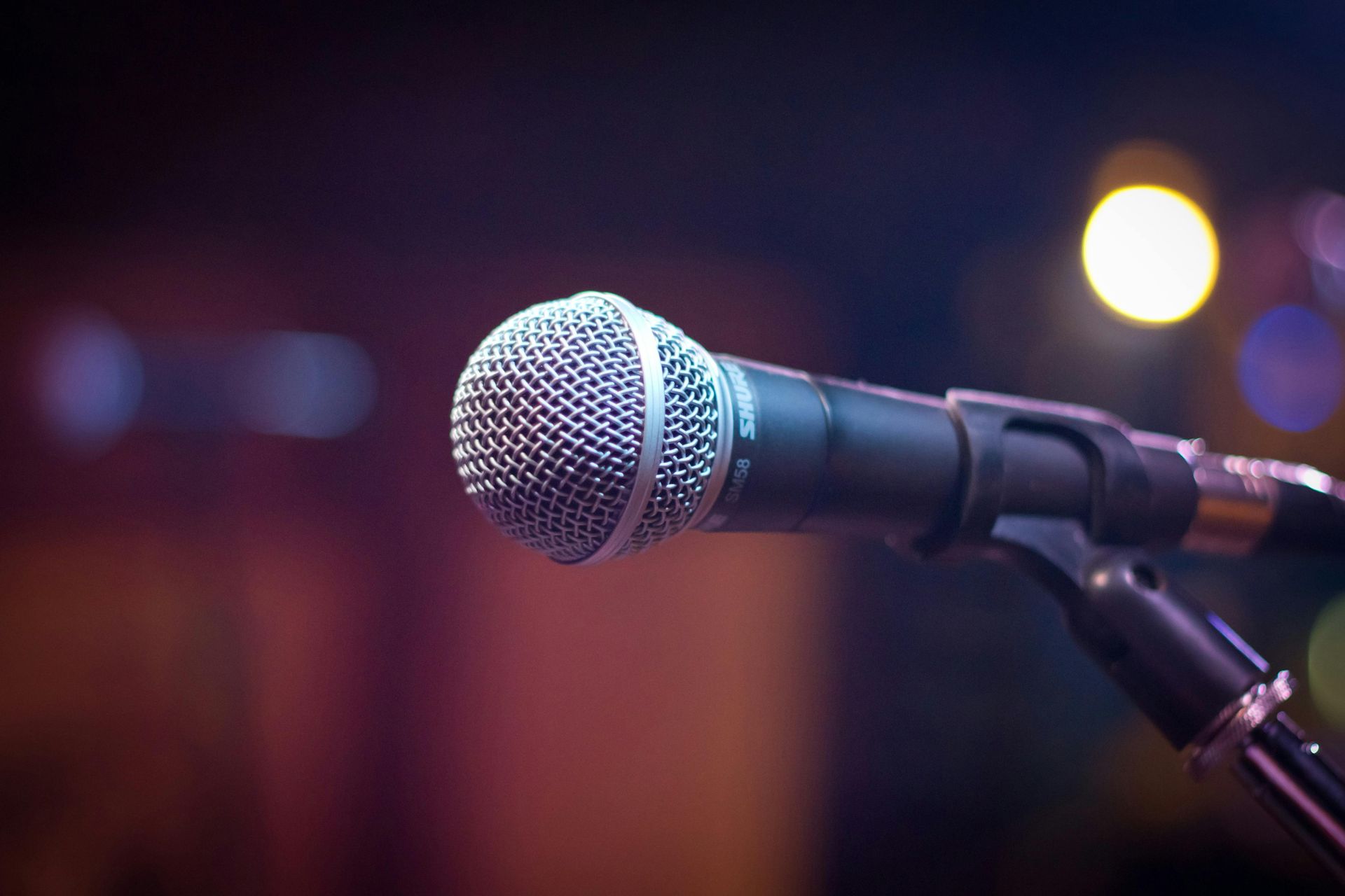 A close-up shot of a silver, handheld microphone mounted on a stand against a blurred, dark stage with warm lighting.