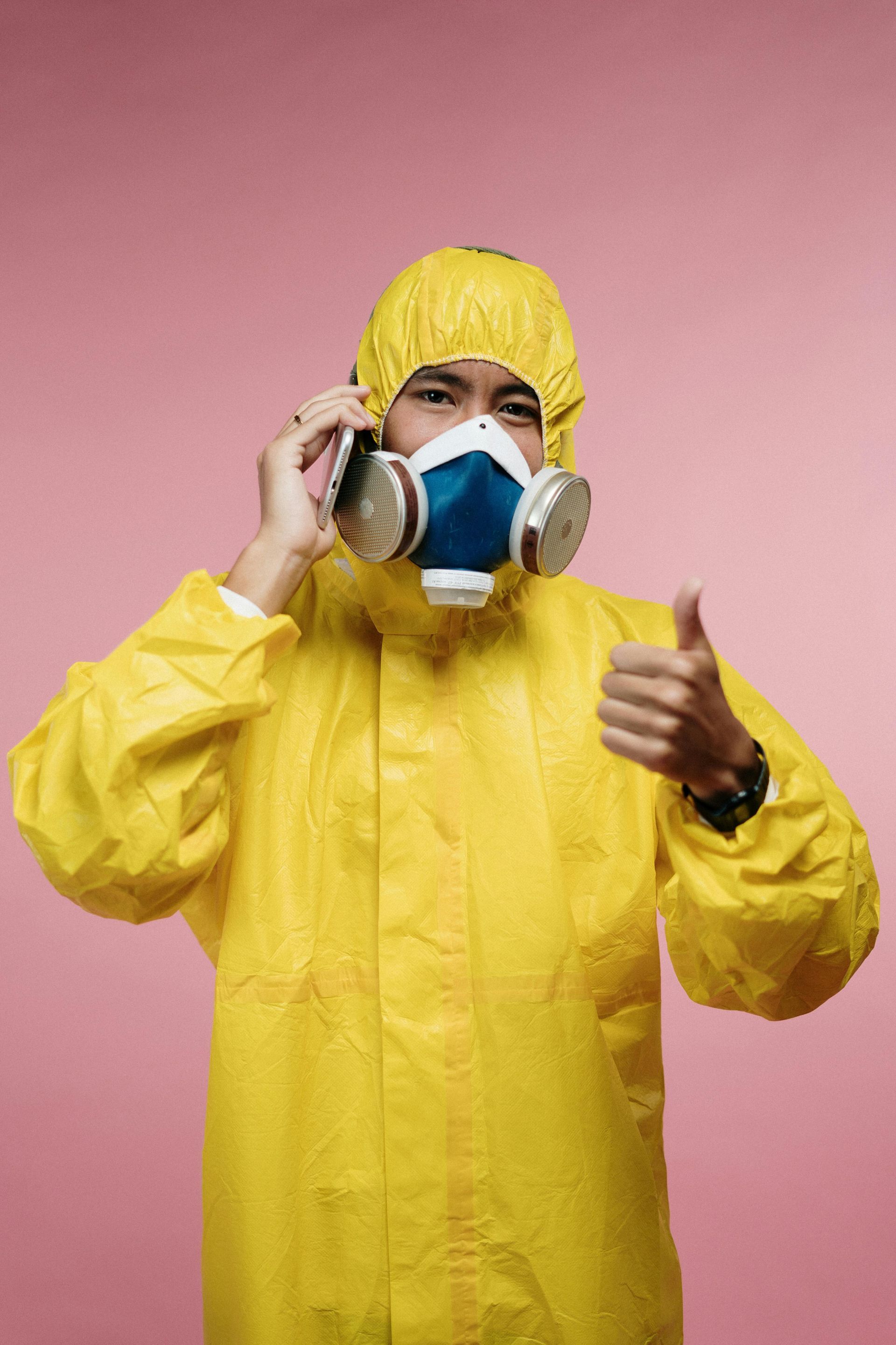 Person in yellow hazmat suit and gas mask on pink background, holding a phone and giving a thumbs-up.
