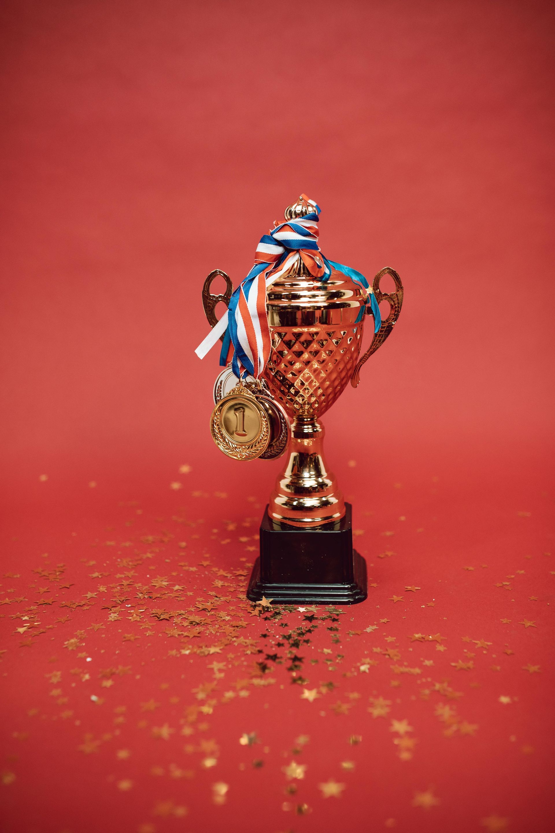A gold trophy with a ribbon and medal attached, resting on a red surface covered with scattered gold glitter.