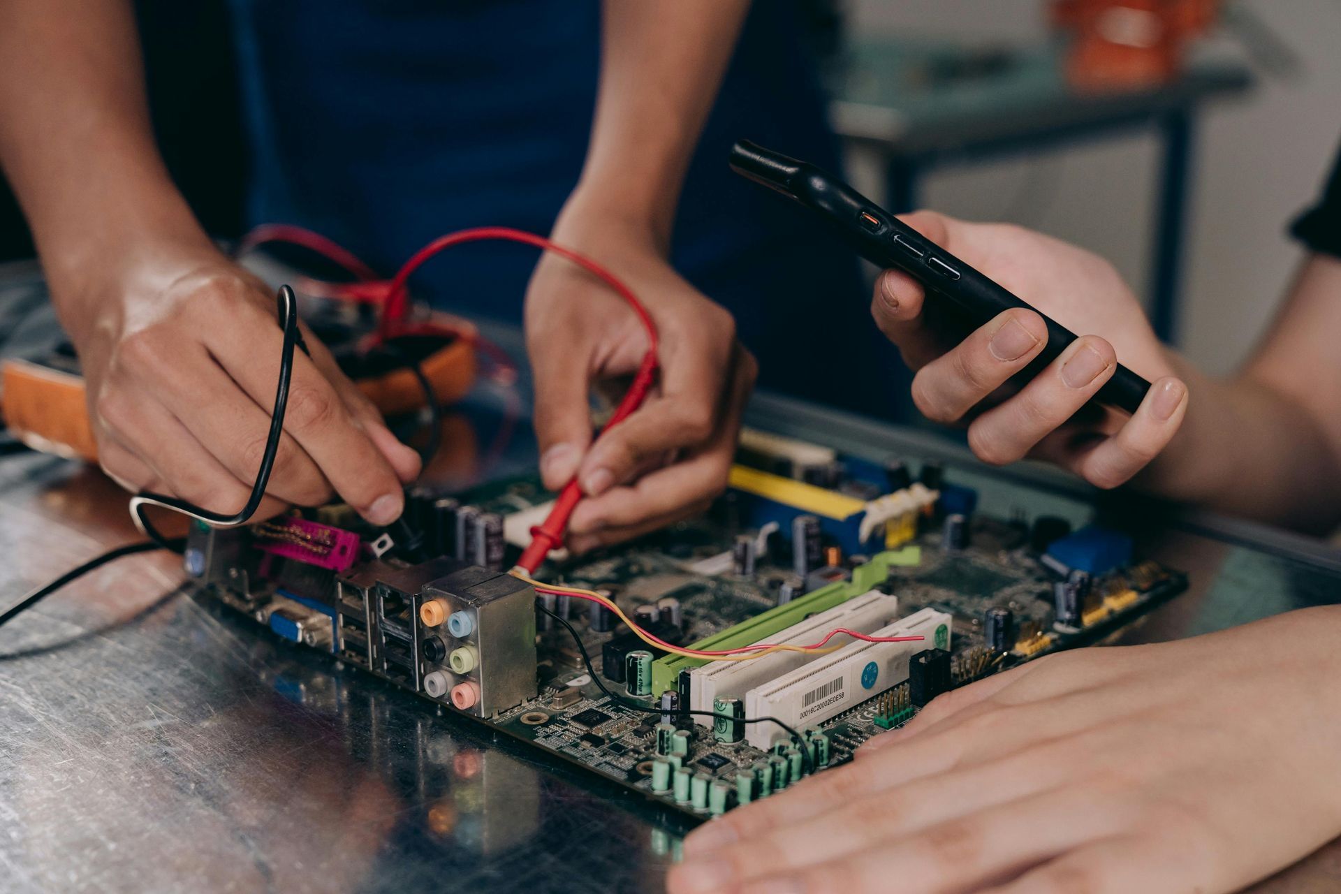 Two pairs of hands work on a computer motherboard with a multimeter and a smartphone.