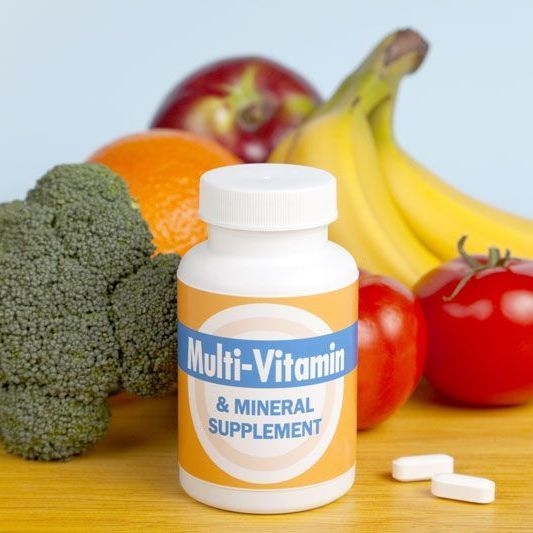 Flacone di integratore multivitaminico con frutta, broccoli e compresse su sfondo chiaro