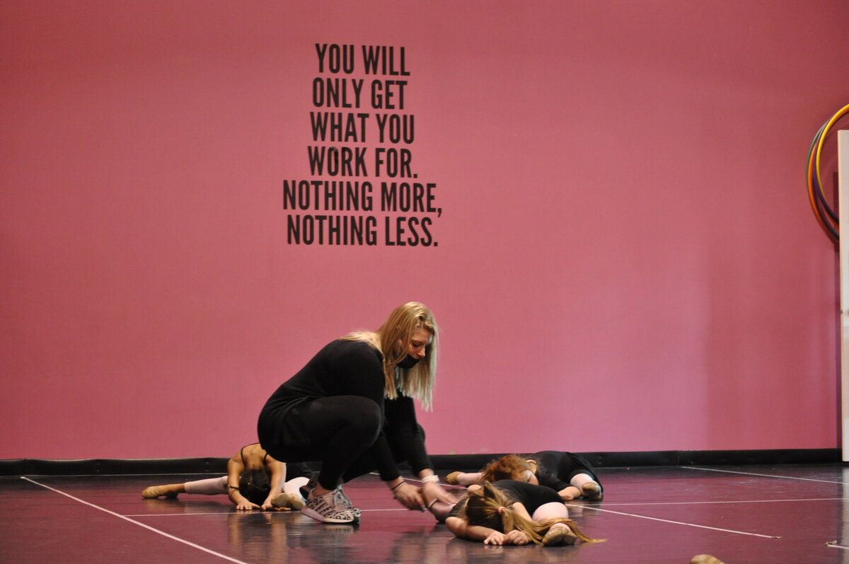 Dancers performing on a pink stage. One kneels to assist two lying on the floor. Quote on wall: