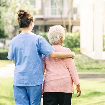 A caregiver in blue scrubs walks outdoors with an arm around an individual in a pink shirt, providing support.