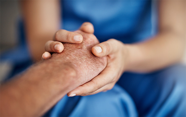 A person in blue scrubs holds the wrinkled hand of another person, providing comfort and support.