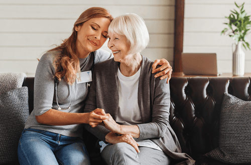 A caregiver embraces and holds the hand of an older person, both smiling gently while sitting on a leather sofa.