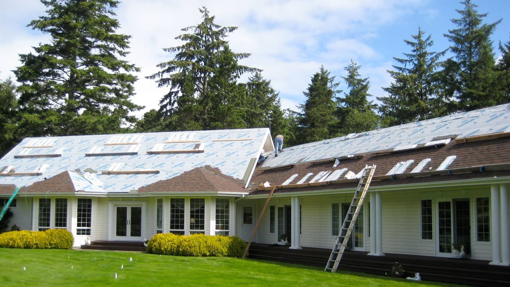 White house with a partially covered roof and ladder on a green lawn, surrounded by tall trees.
