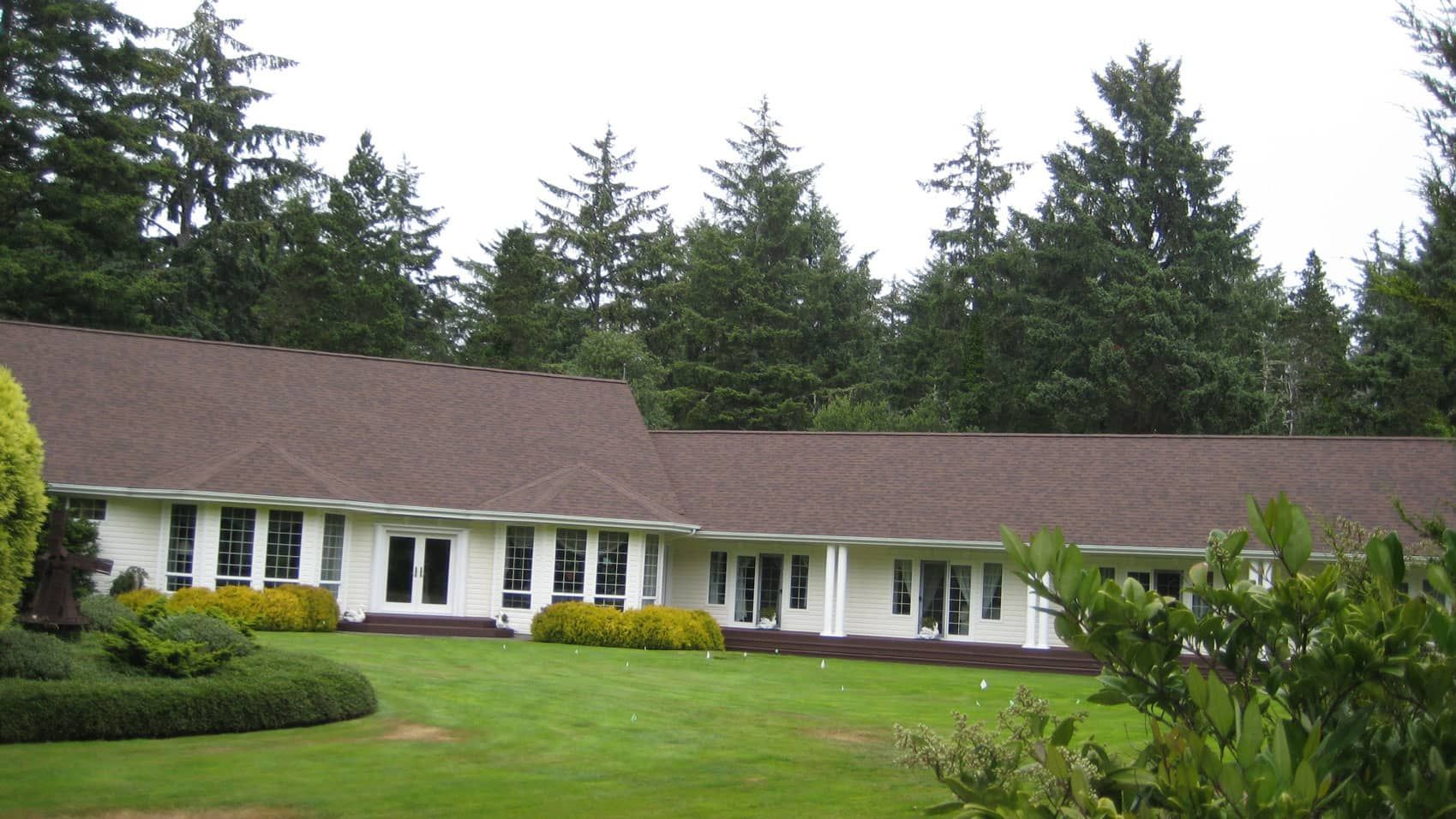 Single-story white house with brown roof and large green lawn, backed by tall evergreen trees