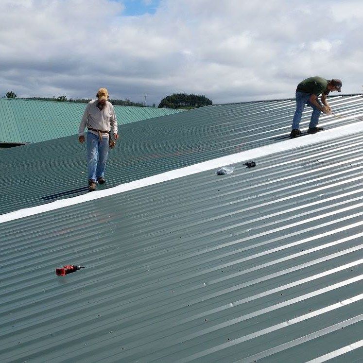 Two workers installing green metal roofing on a large roof under cloudy skies