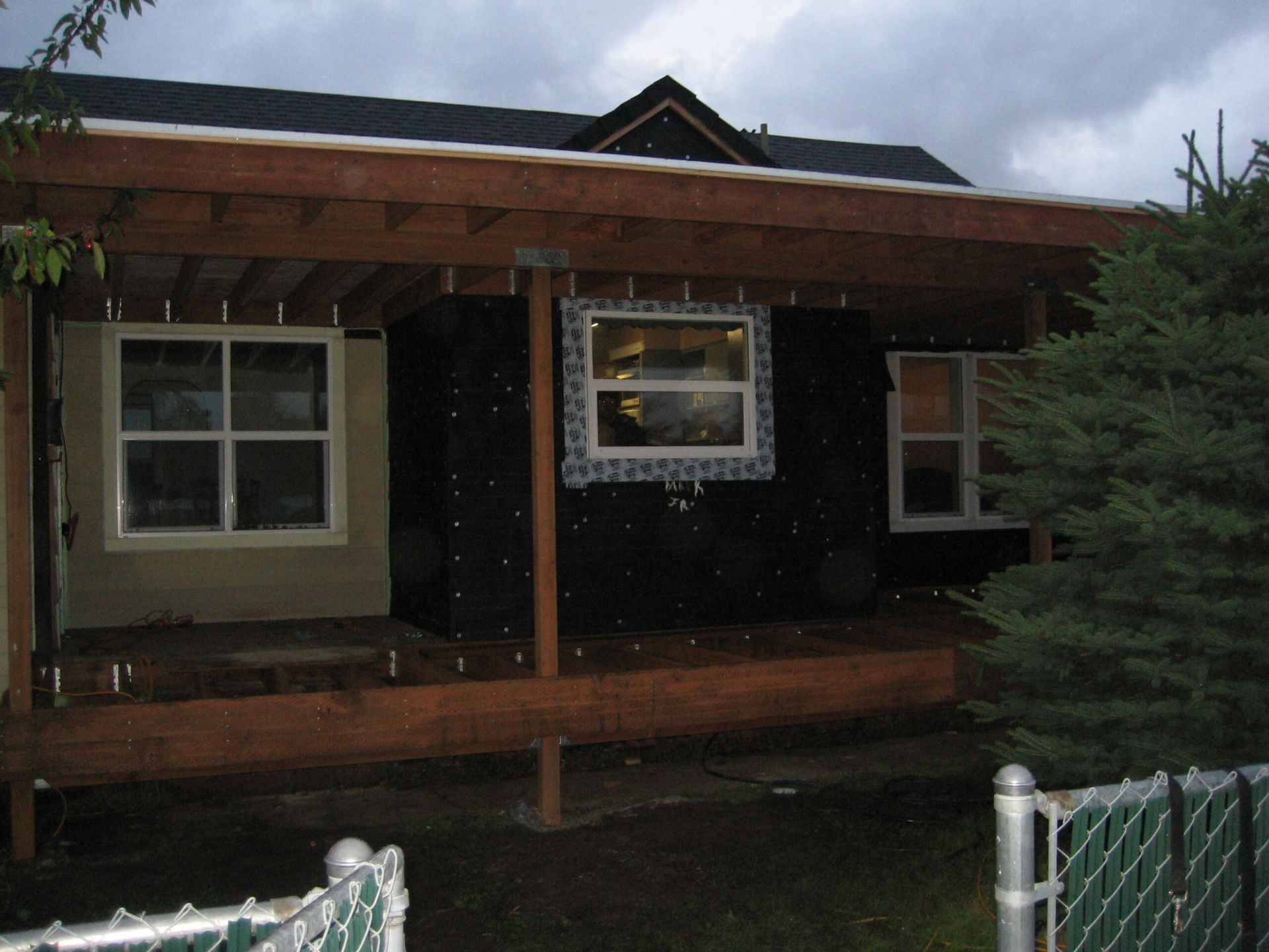 Small dark house with a wooden porch and white-trimmed windows behind a chain-link fence.