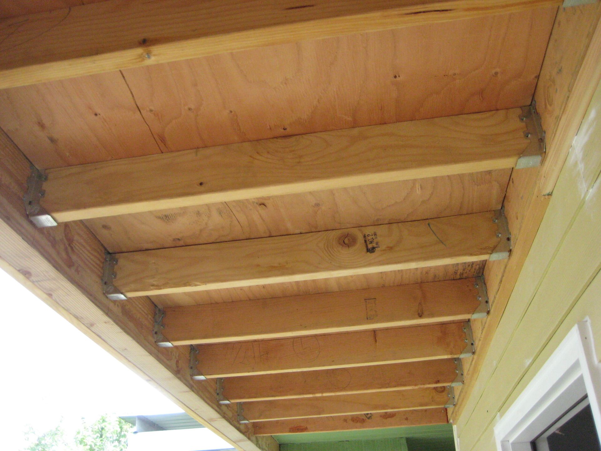 Wooden deck or porch ceiling with exposed beams and joists, viewed from below beside a house wall.