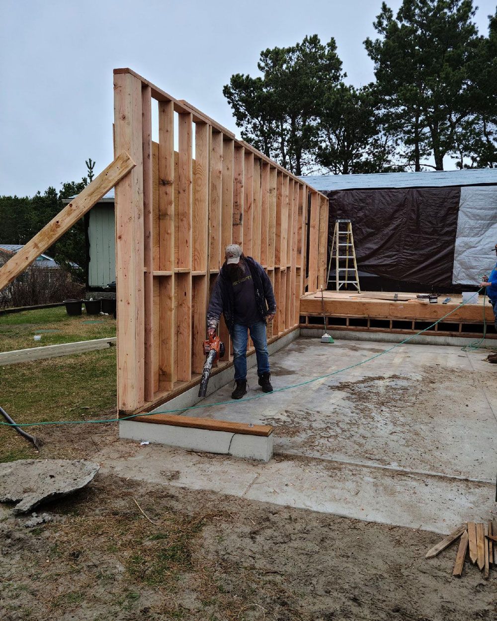 Worker framing a new wooden wall beside a partially built structure on a concrete slab.