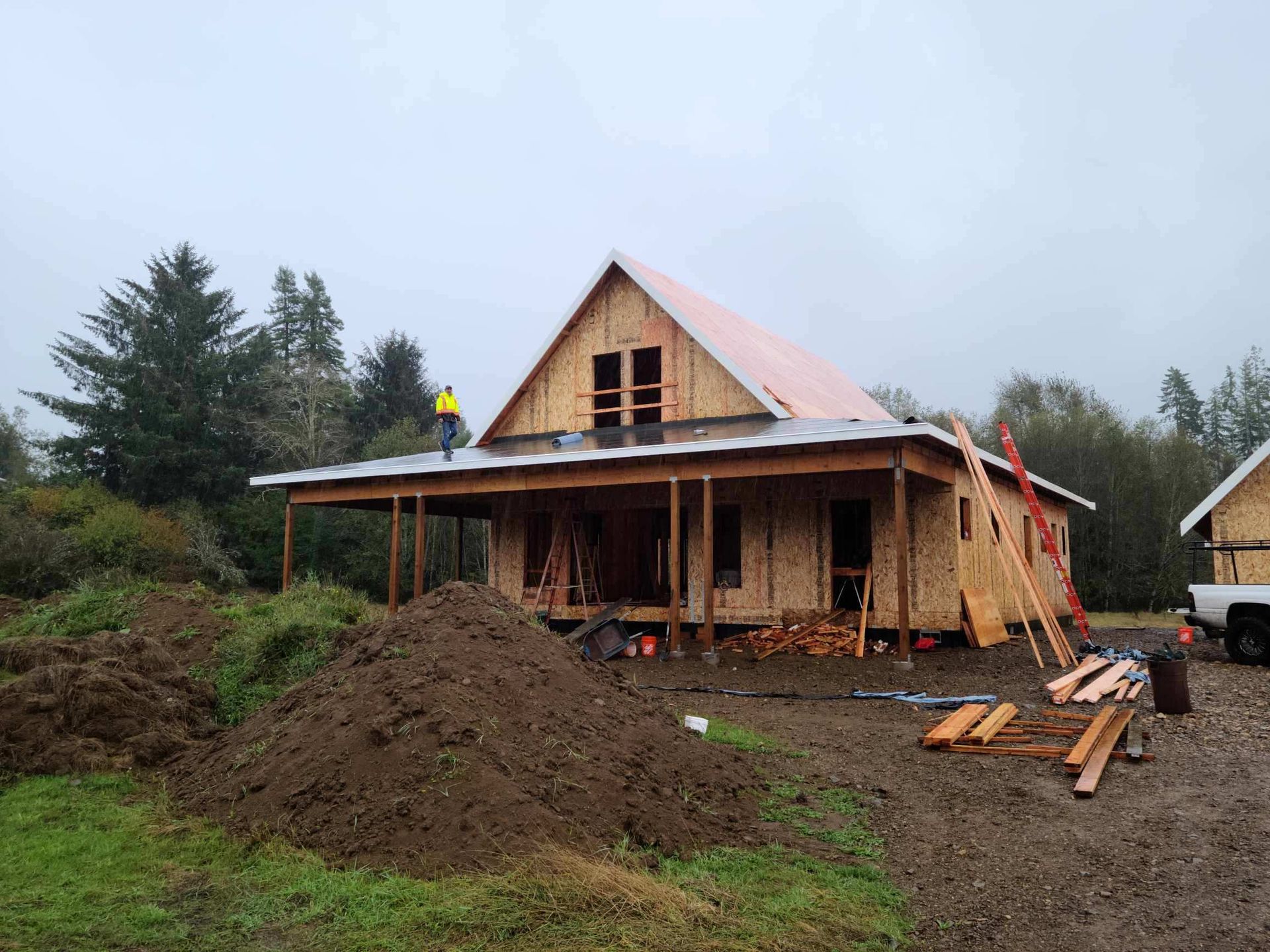 House under construction with workers on the roof and porch, piles of dirt and lumber in a muddy yard