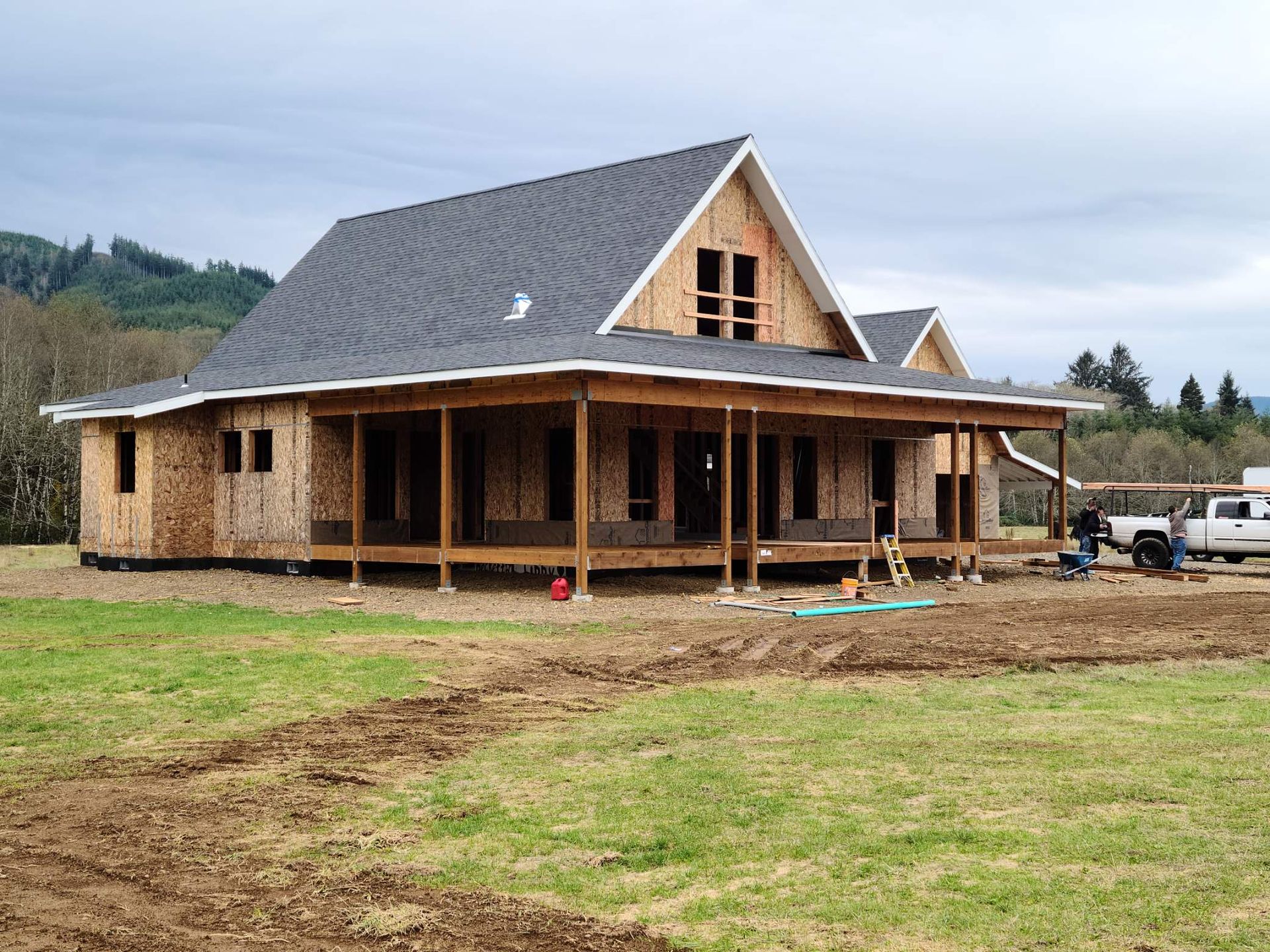 Wood-framed house under construction with a porch and steep gray roof on a grassy lot