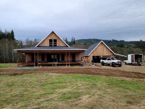 Two unfinished wooden houses with a truck and trailer on a muddy rural lot under a cloudy sky