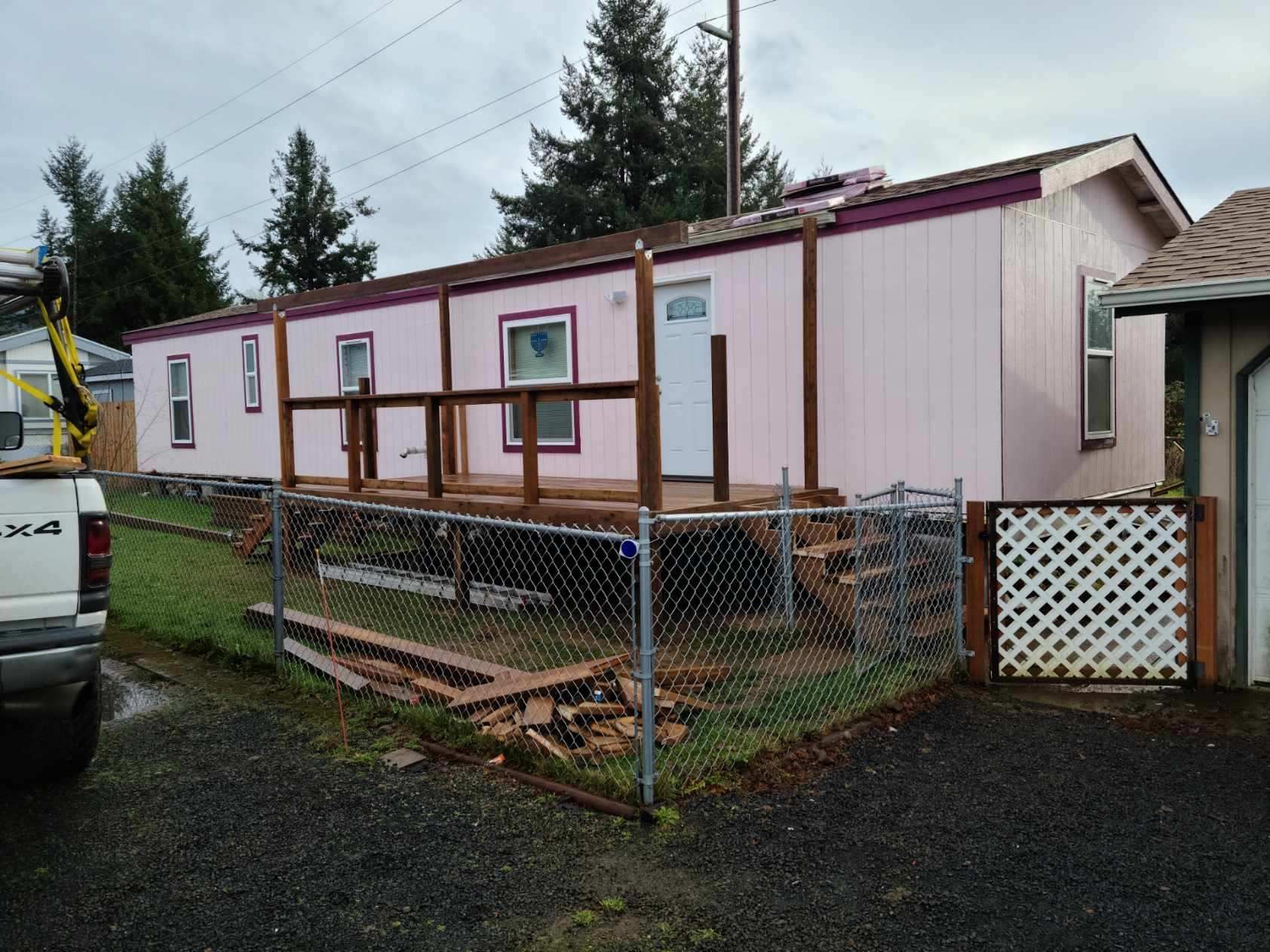 Small pink mobile home with a raised wooden porch and chain-link fence on a rainy residential lot