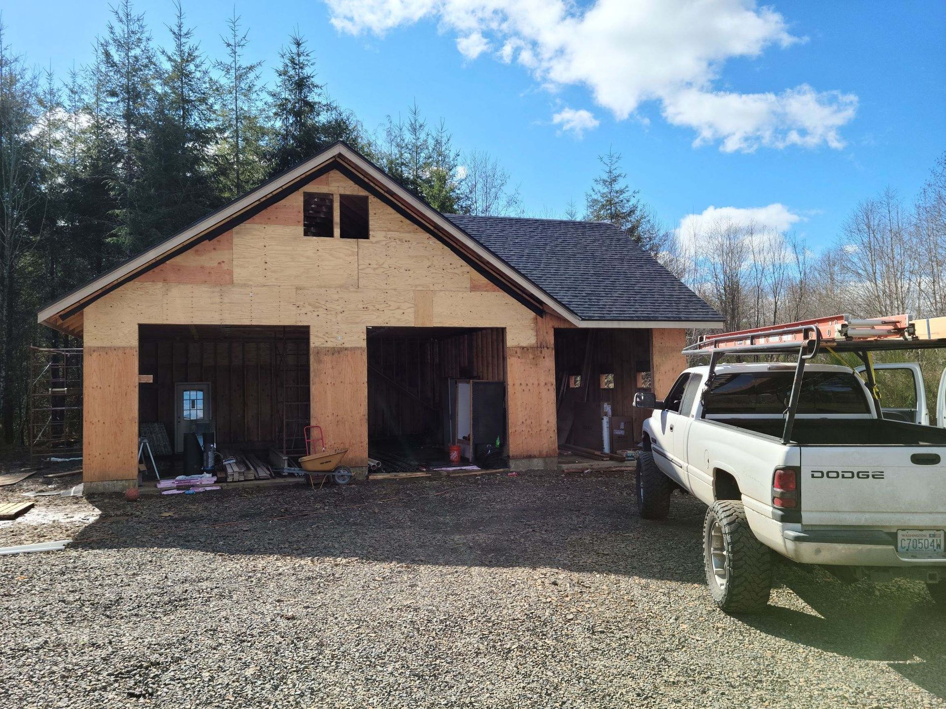 Rural garage under construction beside a white pickup truck on a gravel driveway under a blue sky