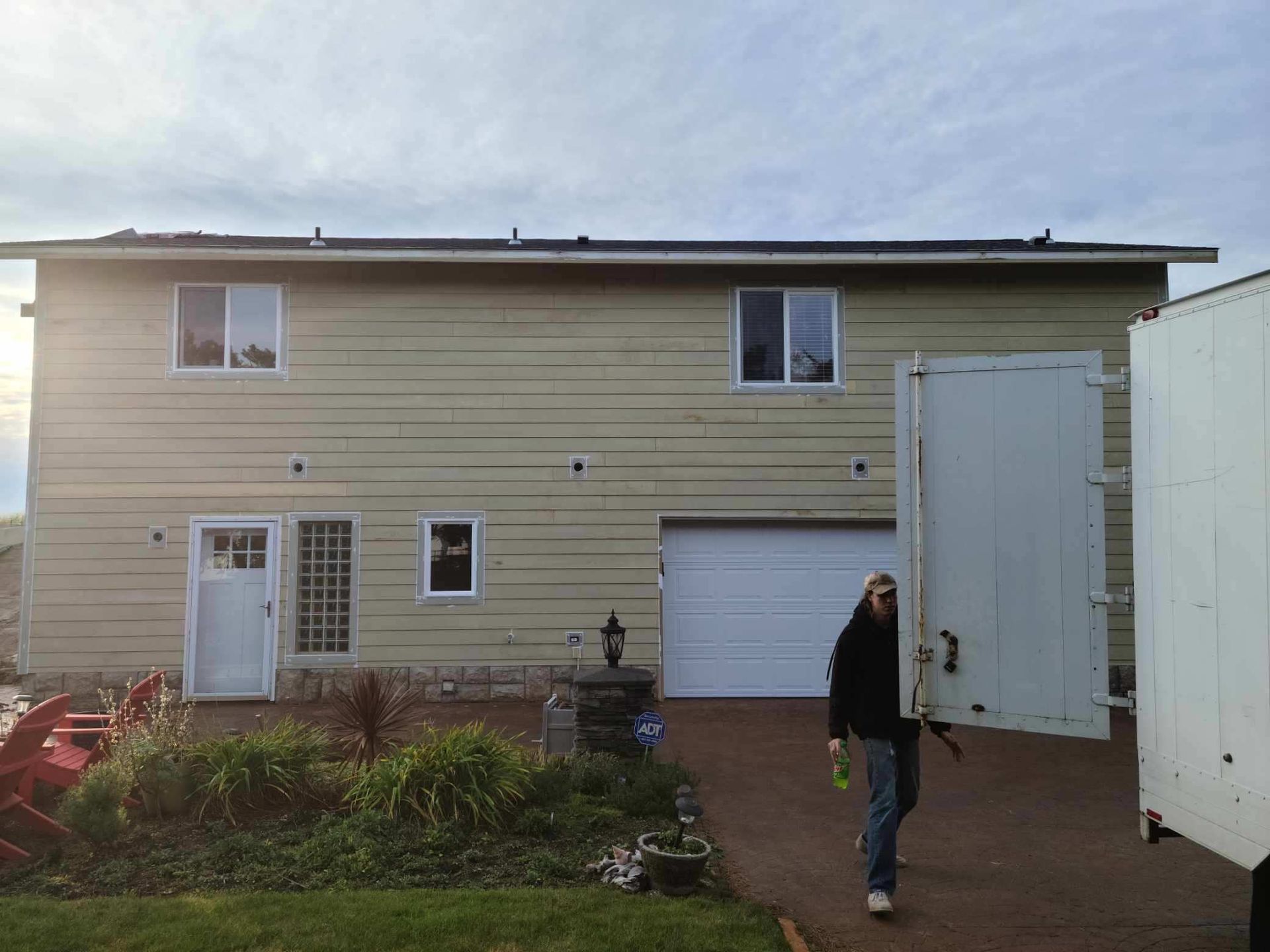 Person opening a large white gate in a driveway beside a beige house with garage and garden bed.