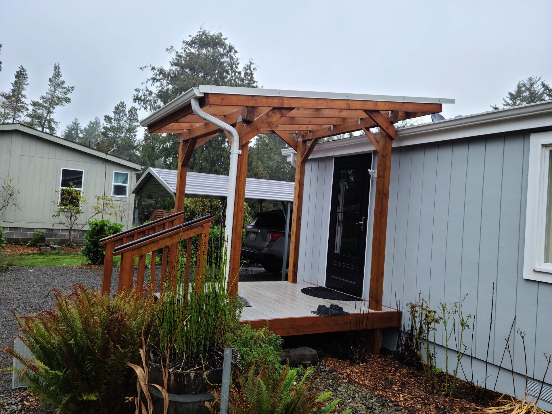 Small wooden porch with stairs and railing beside a gray mobile home in a backyard.