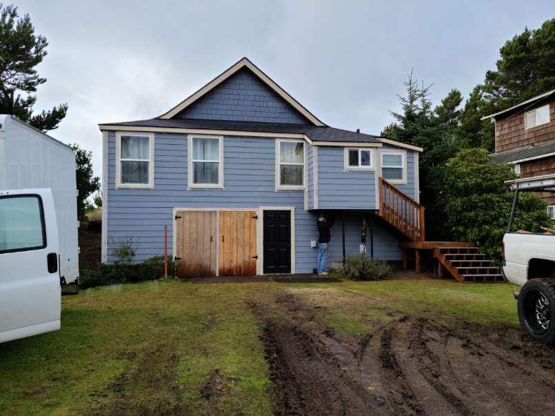 Blue house with a wooden porch, stairs, and muddy driveway, viewed from the front
