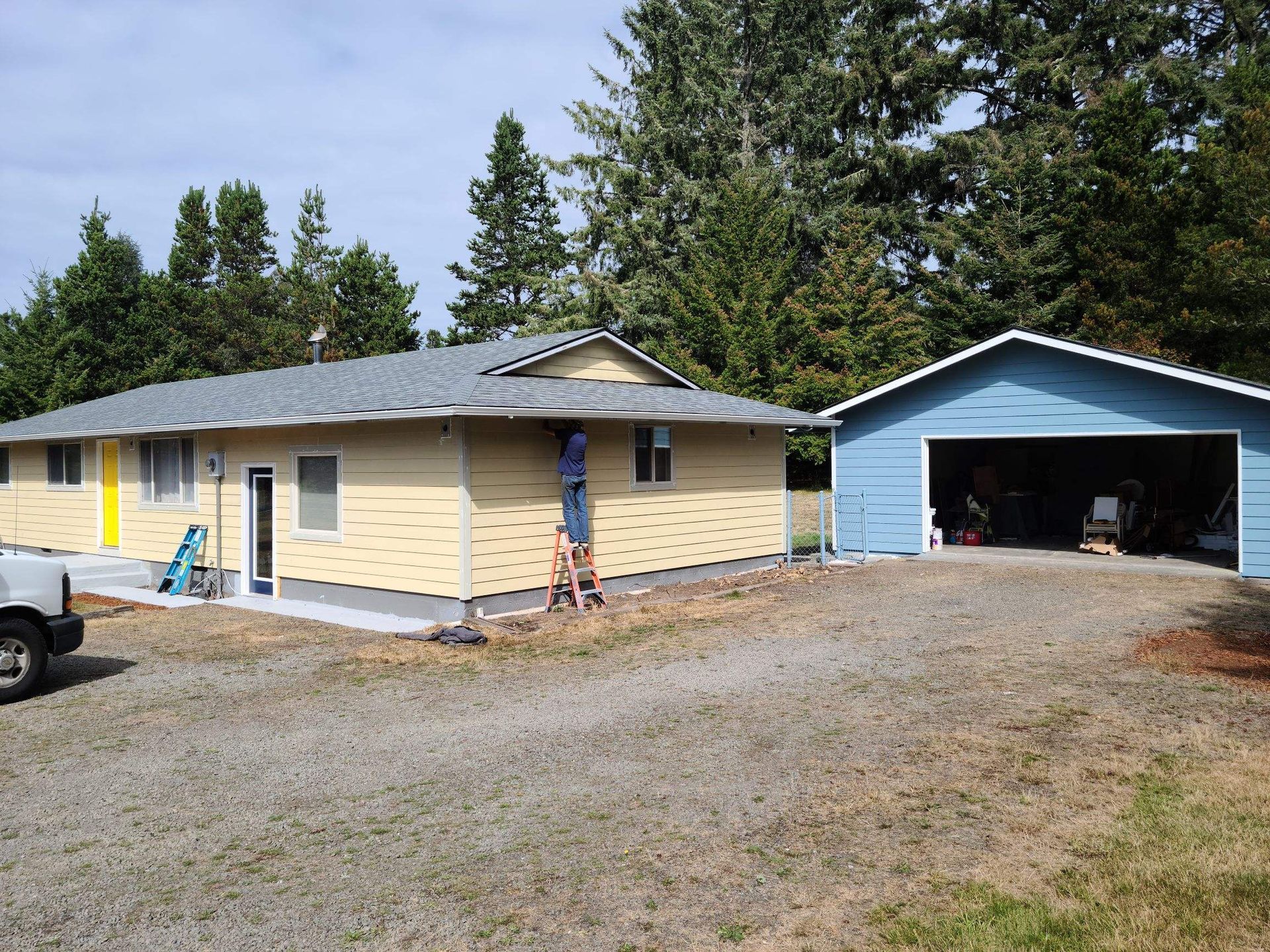 Single-story beige house with blue garage and gravel driveway under trees