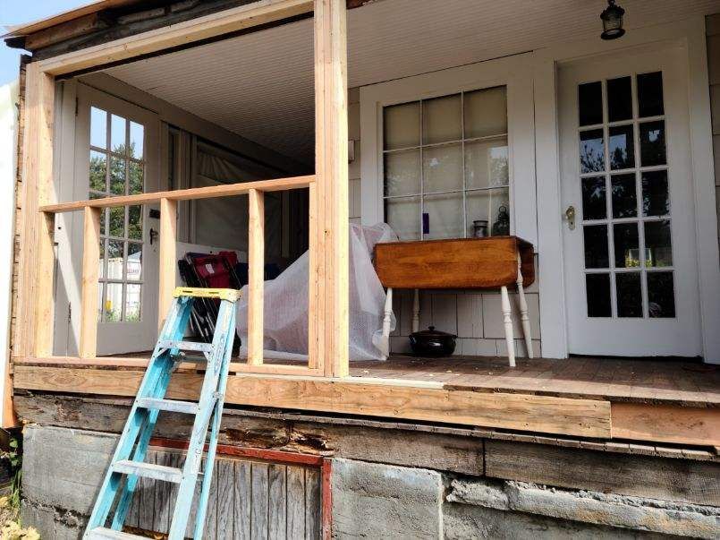 Porch under renovation with unfinished railing, blue ladder, and white front door.