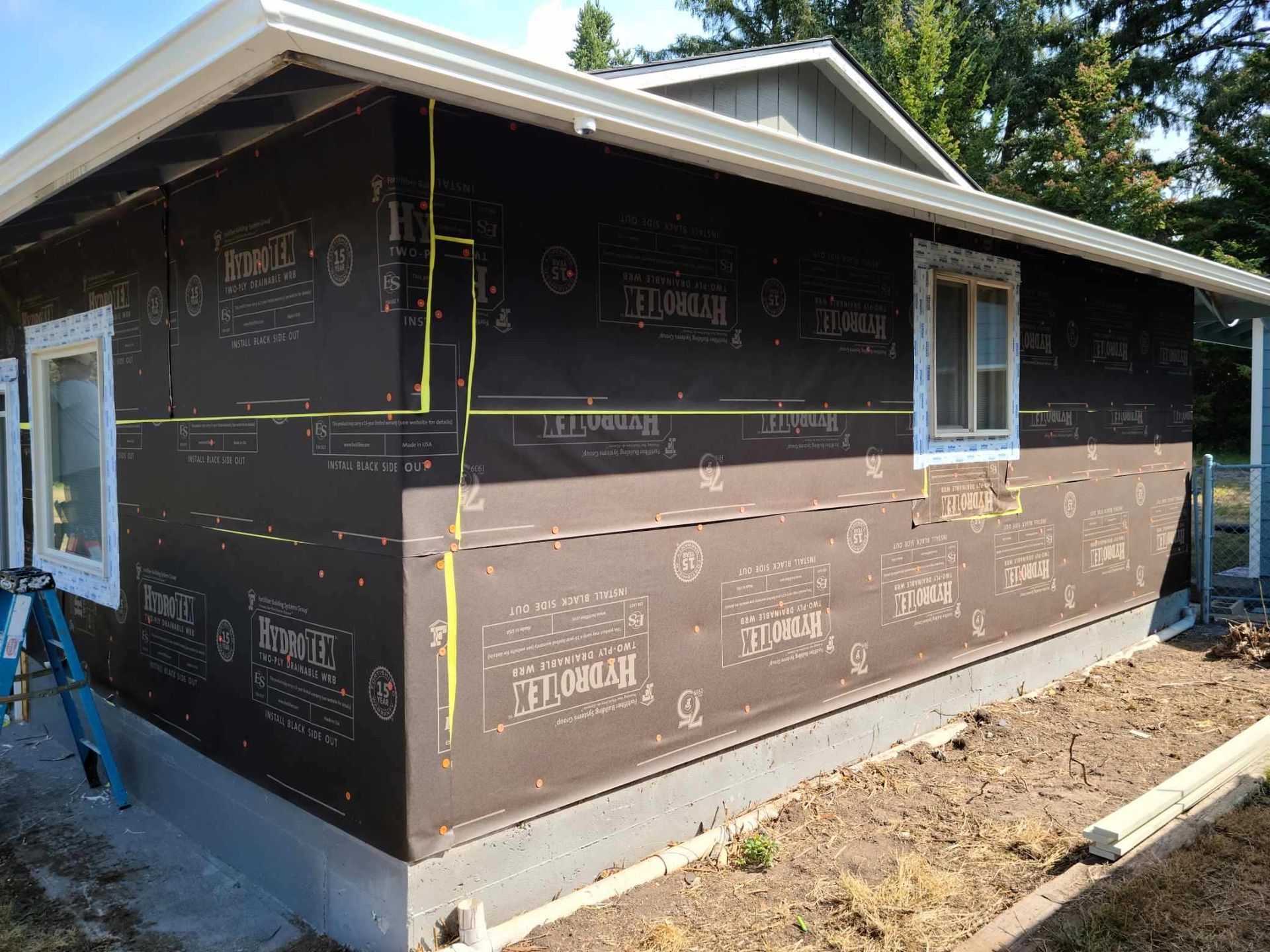 House under construction wrapped in black house wrap, with tan siding and white-trimmed windows