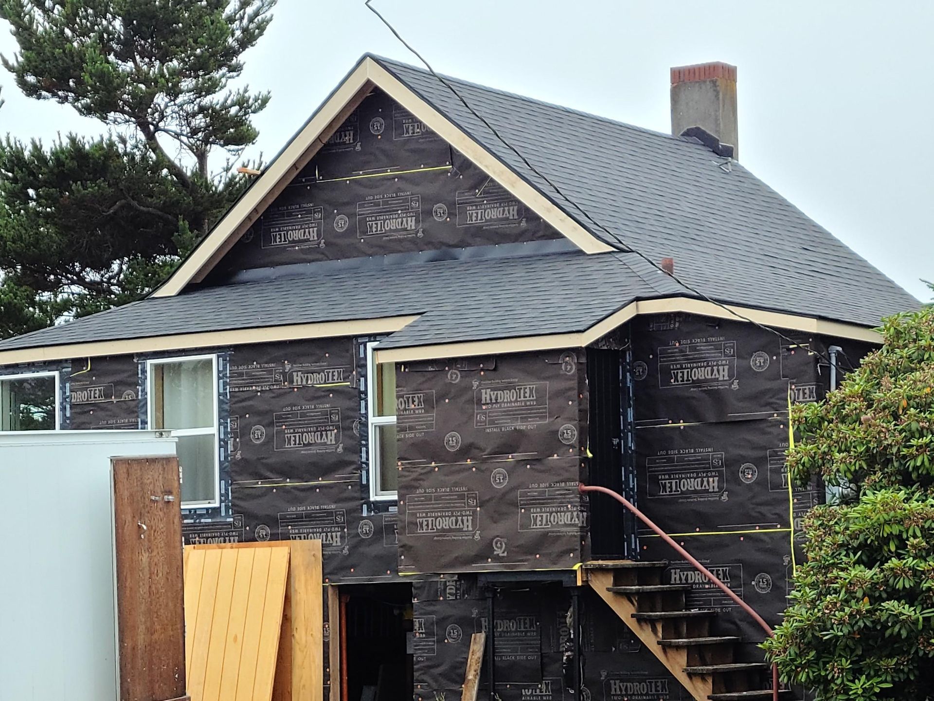 Black house with a steep gray roof and exterior stairs, shown during renovation with bare plywood siding.