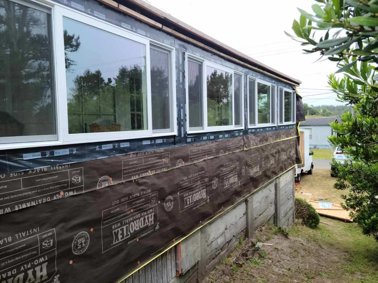 Partially built house exterior with wrapped walls, large windows, and a grassy yard beside a driveway