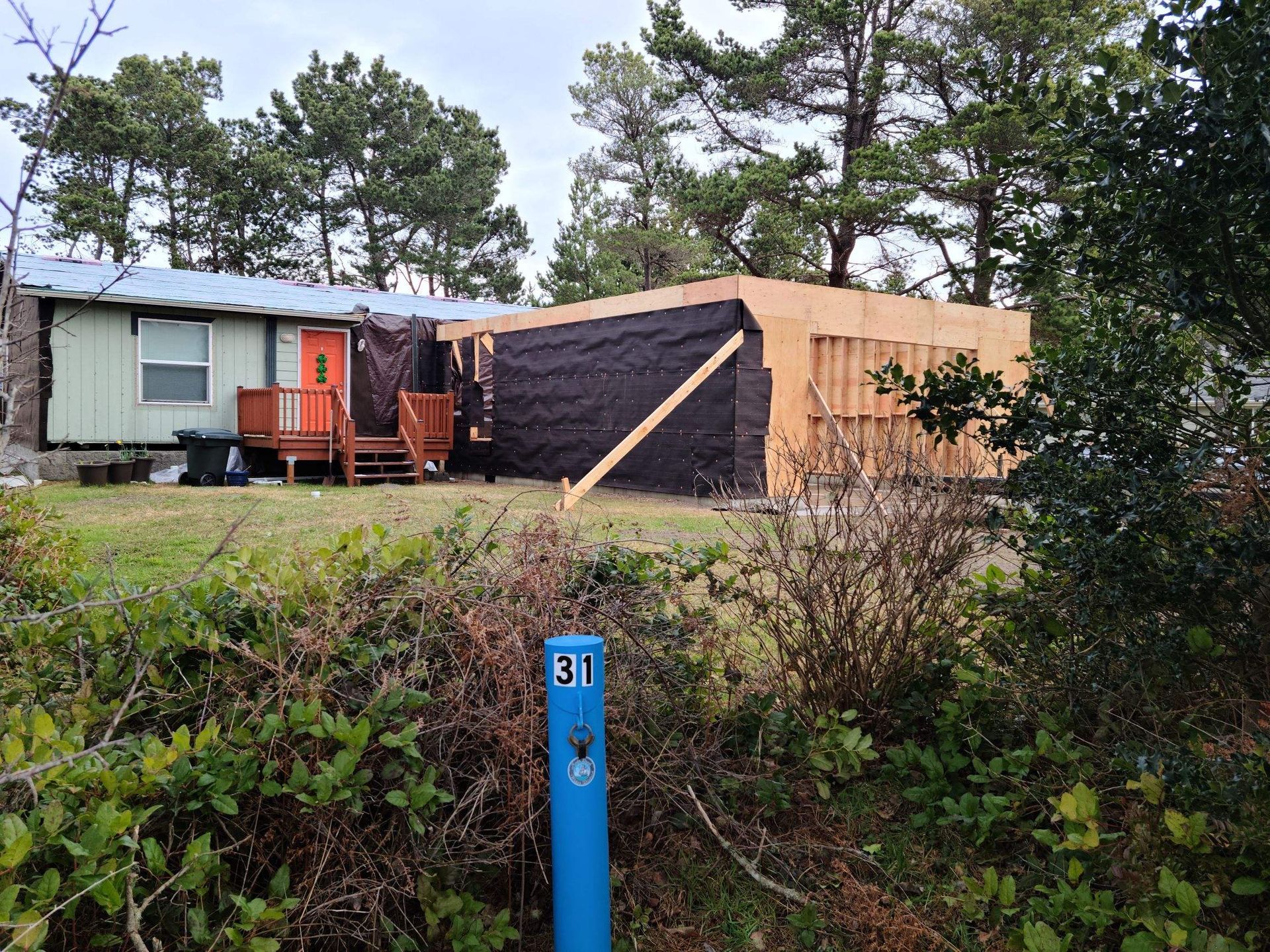 Small house with orange deck beside a large black-and-wood structure, seen behind a blue marker post and brush.