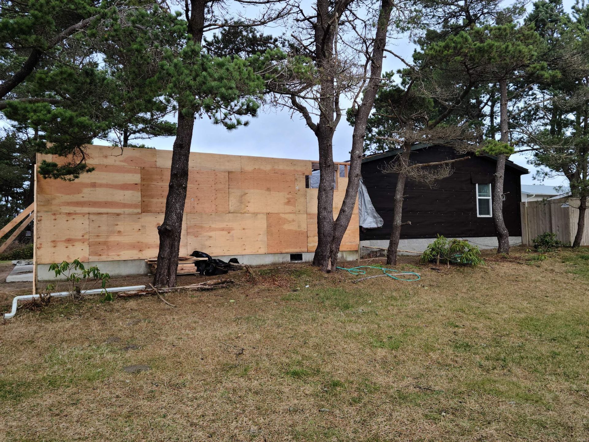 Plywood-covered wall beside a small dark building under trees on a grassy lot