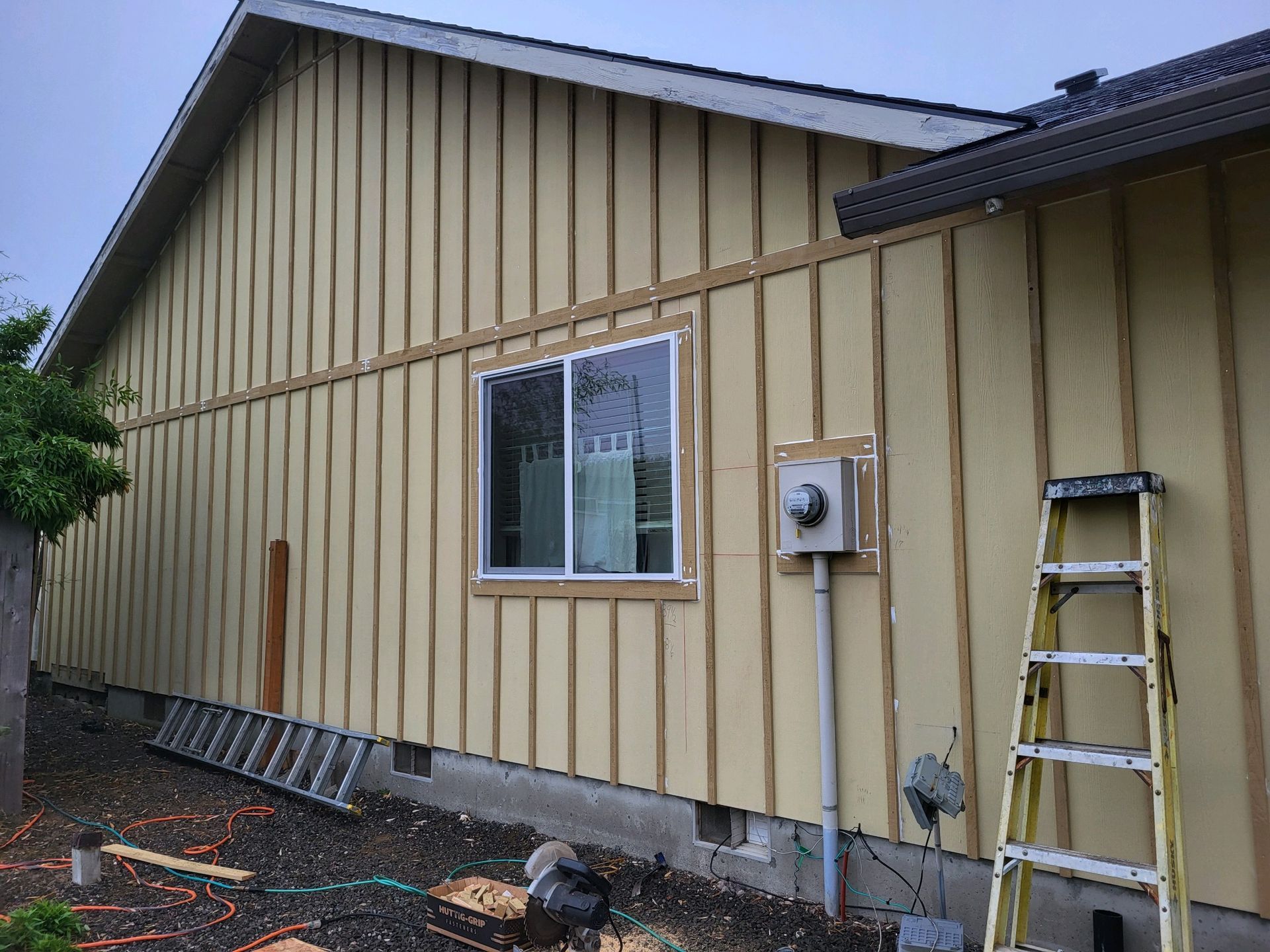 Exterior side of a beige house with new siding, a window, an electrical meter, and a ladder leaning against the wall