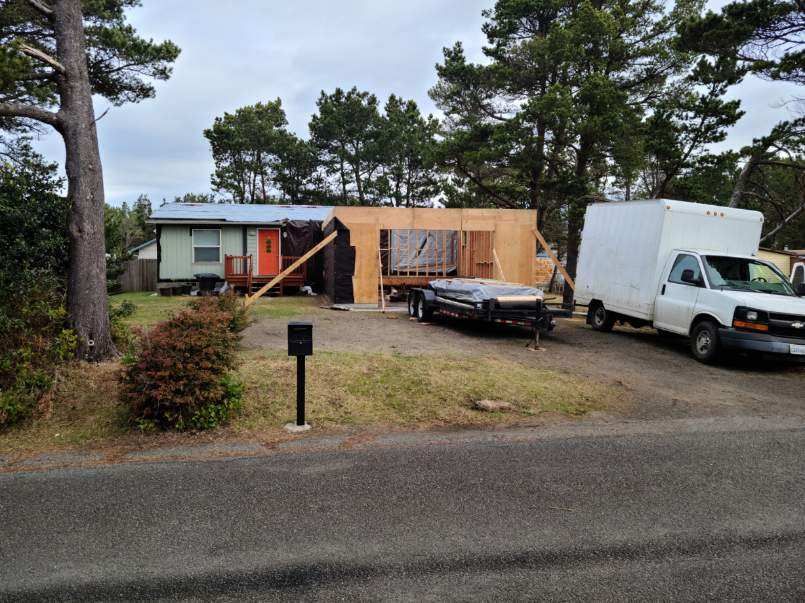 House under construction with exposed framing, a car and white van parked outside on a roadside lot.