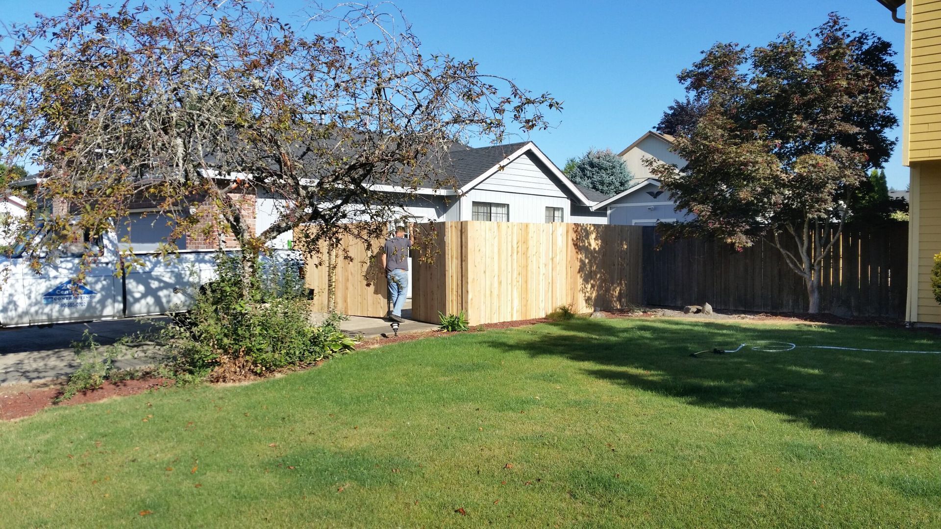 Sunny backyard with green lawn, wooden fence, and a small shed beside a house under clear blue sky