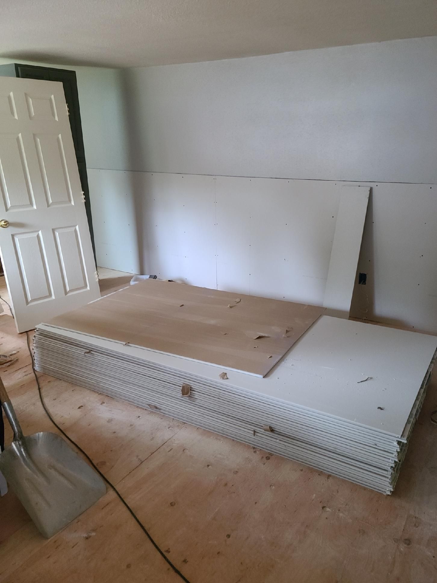 Unfinished room with stacked drywall sheets, plywood, and a partially open door.