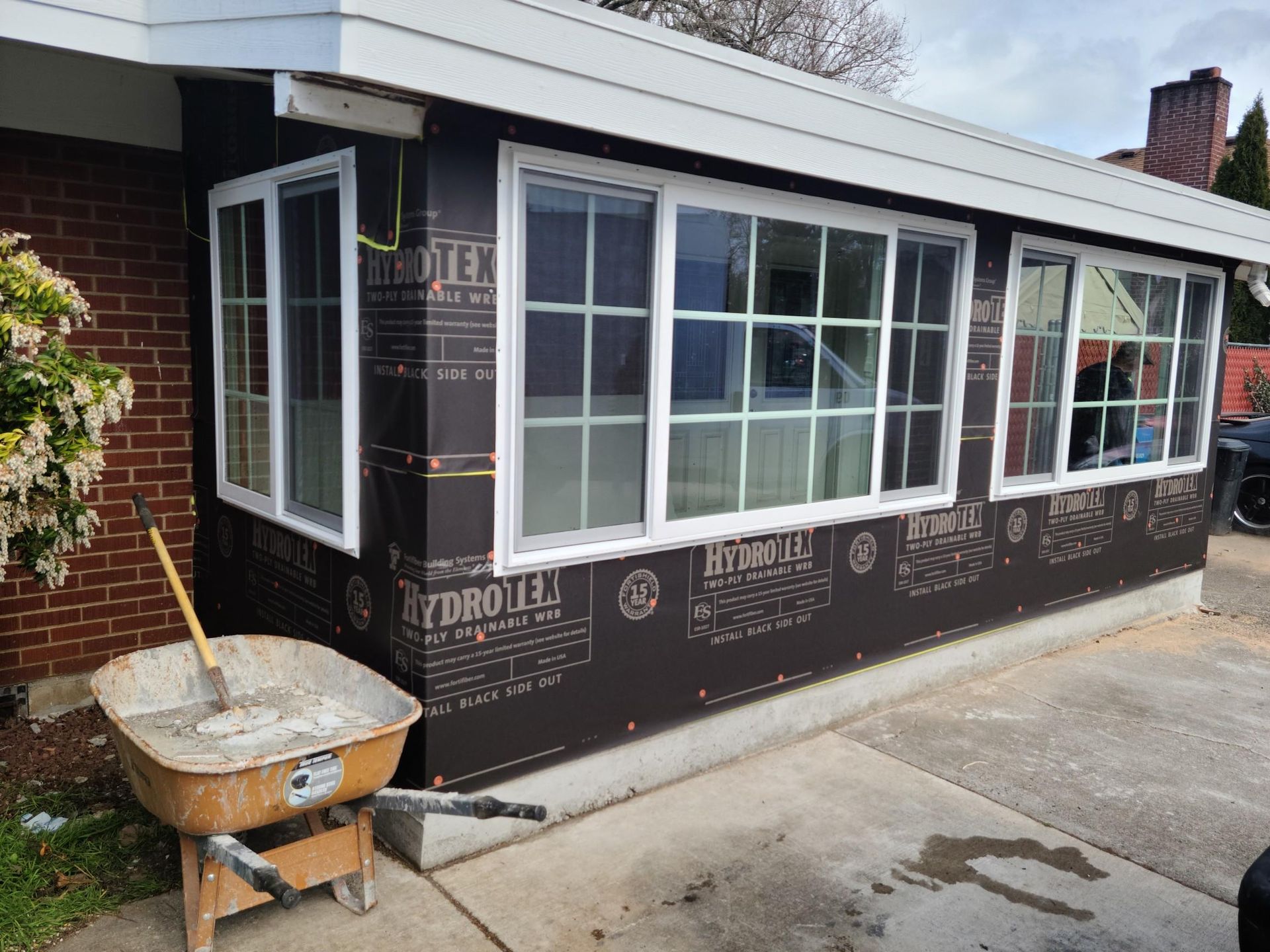 Small brick house addition under construction, wrapped in black weatherproofing with new white-framed windows.