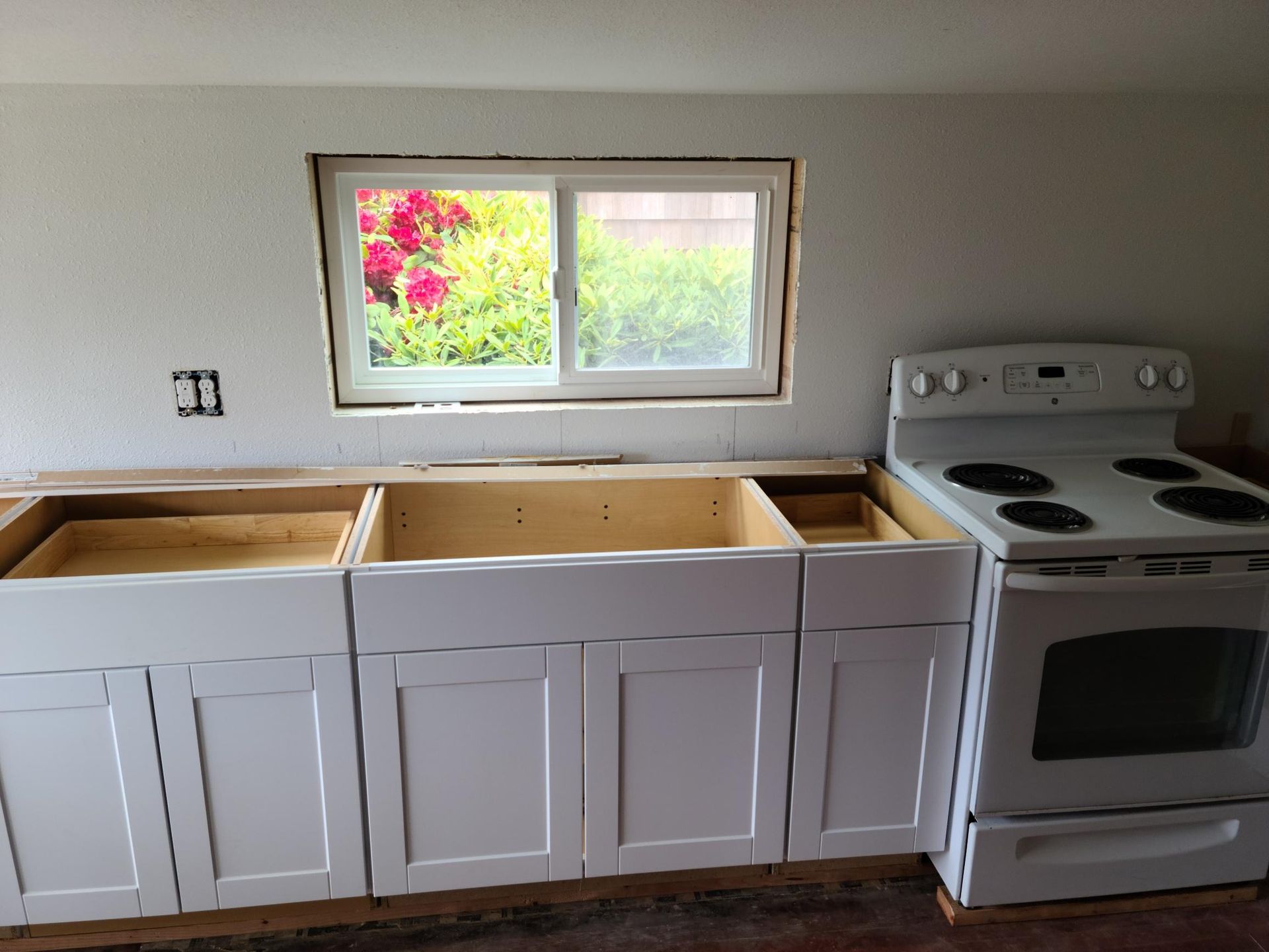 Unfinished kitchen with white cabinets, open sink cutouts, and a white electric stove beneath a window.