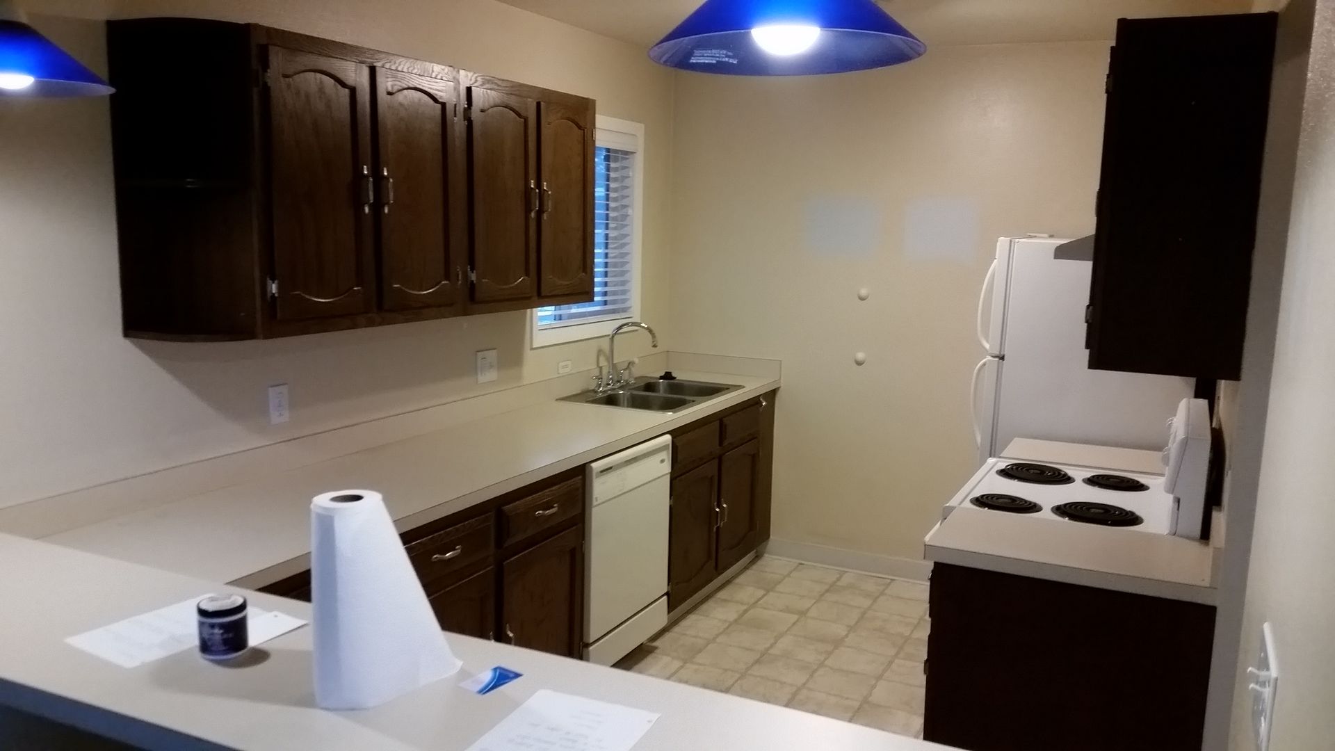 Empty kitchen with dark cabinets, white countertops, and blue pendant lights.