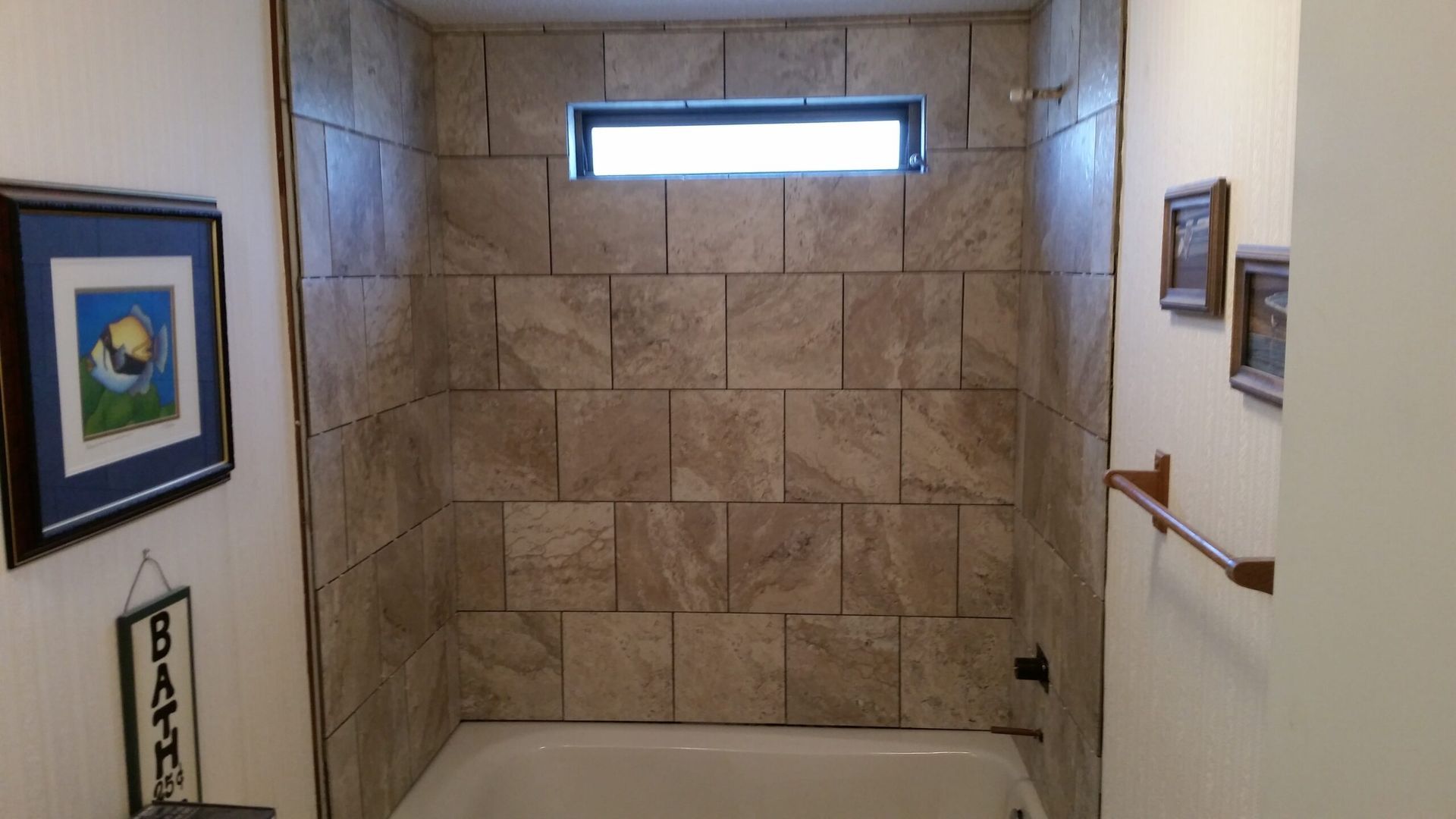 Bathroom tub with tiled shower wall, small window, framed art, and handrail.