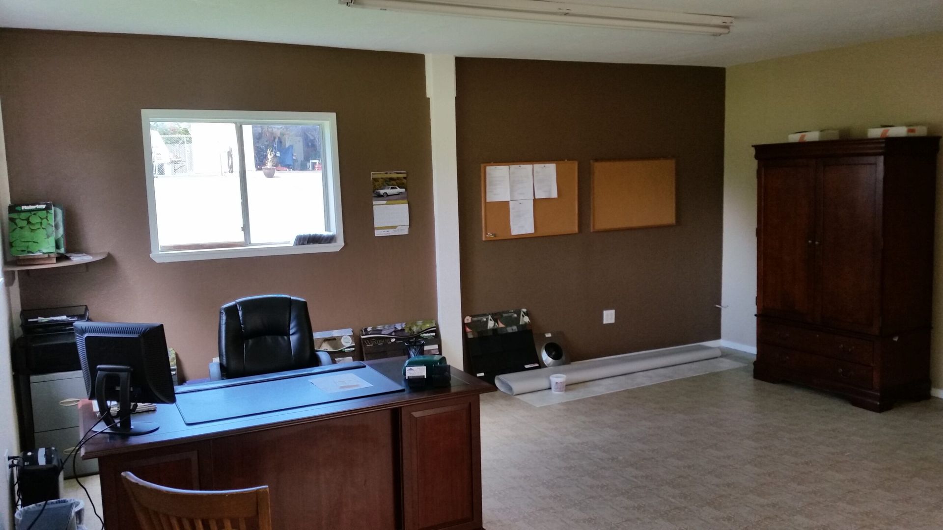 Office room with brown walls, desk, chairs, wall notices, and a large wooden cabinet.