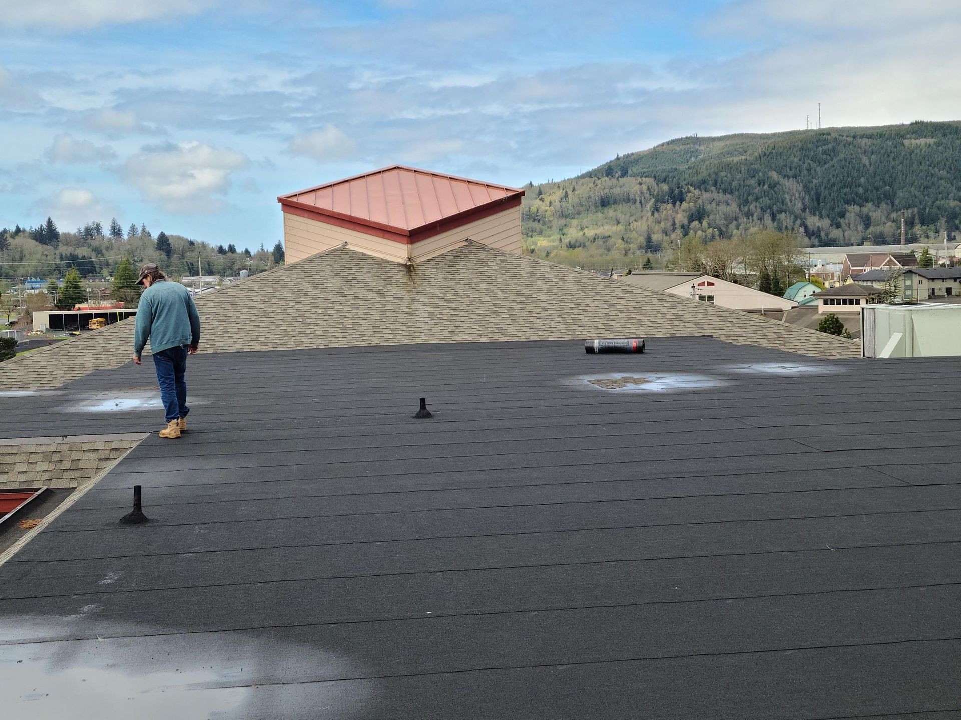 Man walking on a flat black rooftop with a red-roofed building and hills in the background
