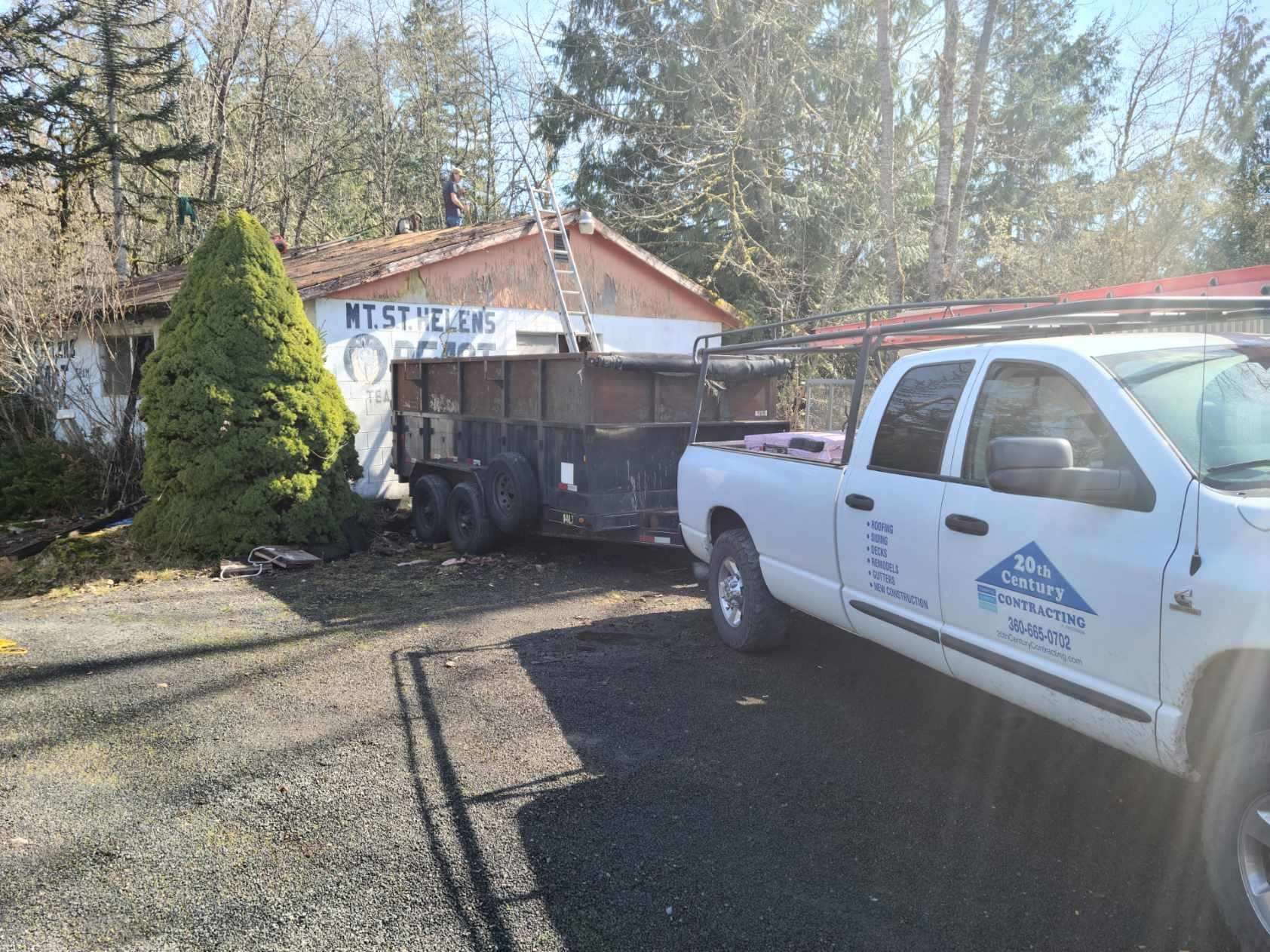 White utility truck beside a black dump trailer in a driveway near a house and trees