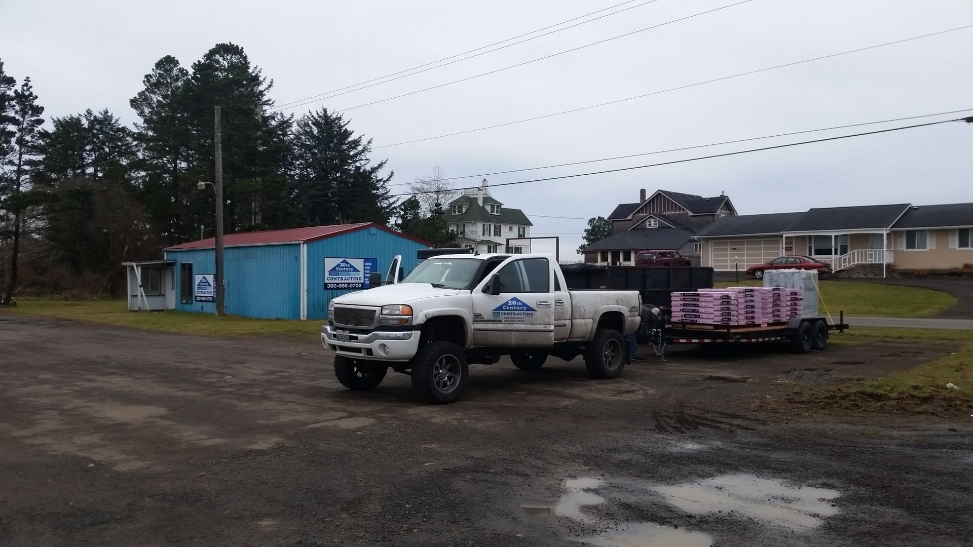 White pickup truck and trailer loaded with pink materials parked on a muddy lot near blue buildings.