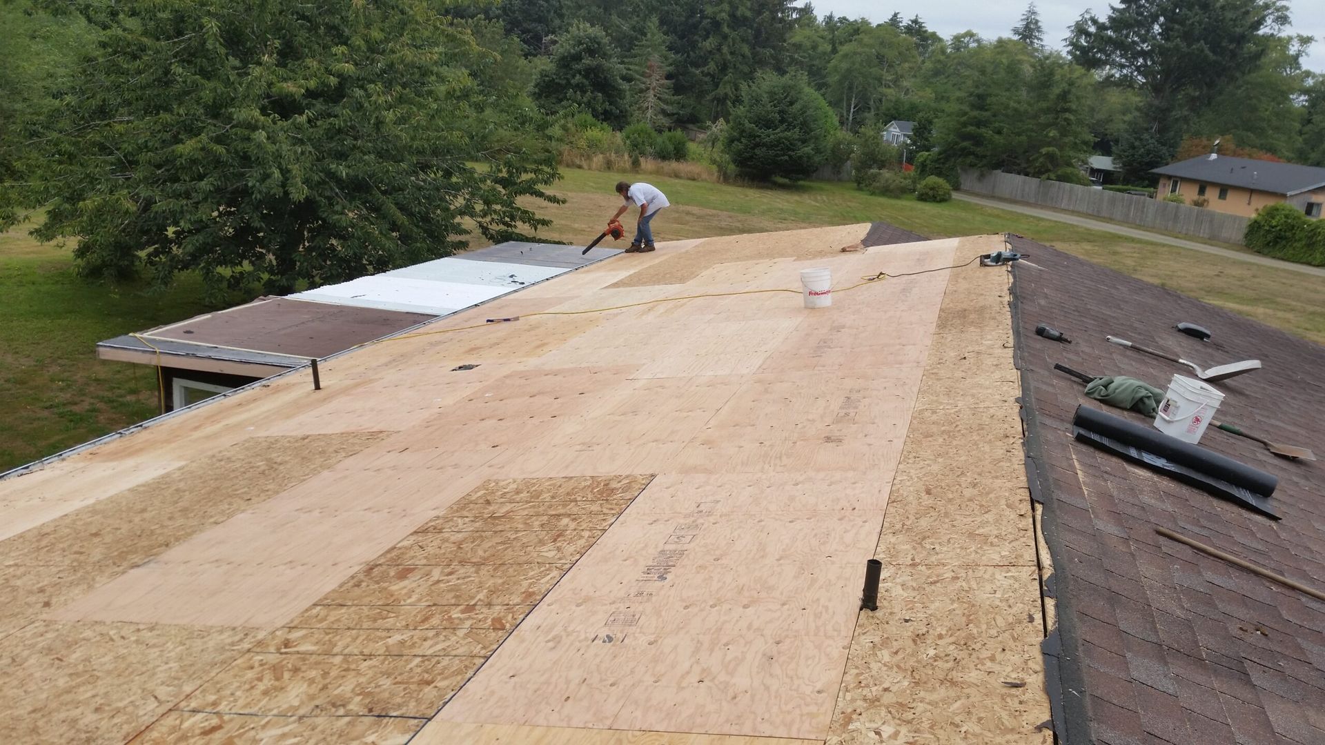 Rooftop under construction with plywood sheathing and workers near trees in the background